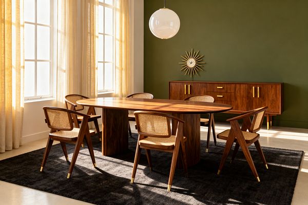 A Mid-Century Modern dining room features an oval walnut table on twin pedestal bases surrounded by walnut armchairs with woven cane seats and backs, all set on a charcoal rug. An olive green accent wall holds a long walnut credenza with brass pulls and tapered legs beneath a brass sunburst clock, while a white globe pendant hangs above and tall windows with ivory linen sheers flood the light-toned floor and walls with warm natural light.