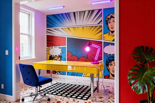 A compact home office framed by red and blue walls features a full comic-book mural with Ben-Day dots across the back wall. A glossy yellow desk with chrome legs sits on a terrazzo floor and black-and-white grid rug, paired with a blue patterned swivel chair. A magenta neon desk lamp and two purple ceiling tubes add glow, while a window brings daylight; a large monstera plant stands at the right.