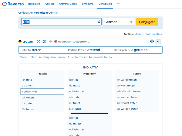 Two young people are sitting outside on a bench, engaged in conversation on a sunny day with greenery and buildings in the background.