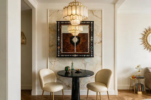A refined studio apartment sitting nook features two cream upholstered chairs around a small black pedestal table, set against a marble-look wall panel with an ornate black mirrored centerpiece. A tiered geometric chandelier hangs above, while gold accents, herringbone wood flooring, and a sunburst mirror add classic Art Deco glamour.