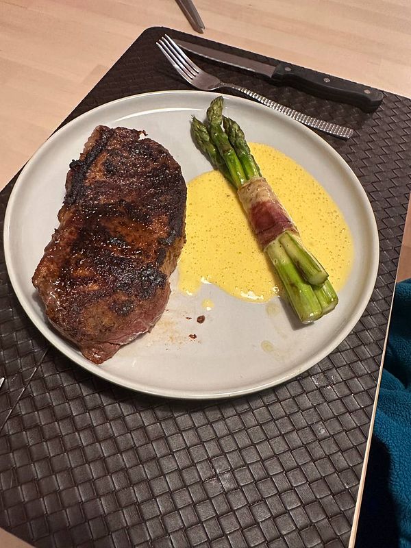 A plate featuring a cooked steak alongside bacon-wrapped asparagus and hollandaise sauce.