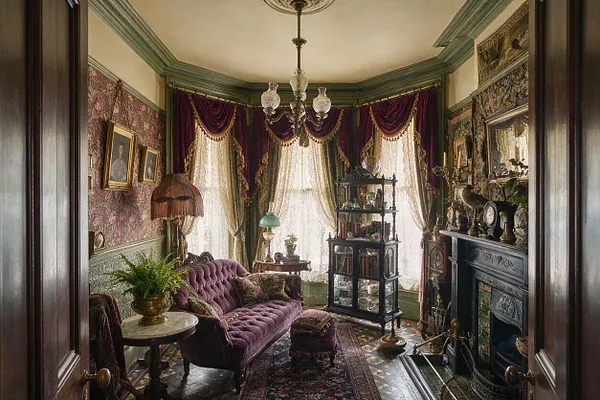 A richly decorated Victorian sunroom with lace-curtained bay windows, deep plum velvet drapes, and patterned wallpaper. The room includes a tufted purple sofa, ornate wood and glass display cabinet, dark carved fireplace, brass accents, and layered rugs under a vintage chandelier.