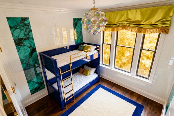 A kids’ room in Hollywood Regency style features a glossy cobalt blue bunk bed with polished brass ladder and rails against white walls inset with emerald malachite panels, lit by an iridescent bubble-glass chandelier. Tall windows with chartreuse satin valance and gold fringe overlook dark hardwood floors and an ivory rug framed by a royal-blue border.