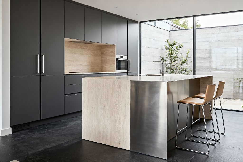 A minimalist kitchen features matte charcoal flat-front cabinetry with discreet stainless handles, a built-in stainless wall oven, and a warm travertine stone backsplash and countertop niche. A sculptural island pairs a travertine waterfall end with a brushed stainless steel counter and integrated sink, accompanied by two tan leather bar stools; dark large-format stone flooring and full-height black-framed windows bring generous natural light and courtyard views.