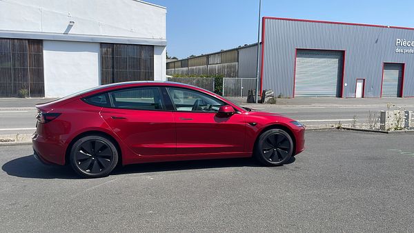A red Tesla Model 3 parked in a driveway with a silver Tesla Model S and a garage in the background.