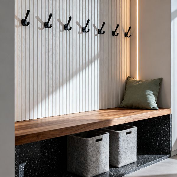 A compact modern mudroom nook features a natural wood bench above a black speckled terrazzo base holding two gray felt storage bins. A white vertical fluted backboard displays a row of matte black hooks, with a slim warm LED light strip illuminating one side; a soft sage green pillow sits at the corner. Daylight casts diagonal shadows across the paneling, highlighting the clean lines, textures, and tidy, functional layout.
