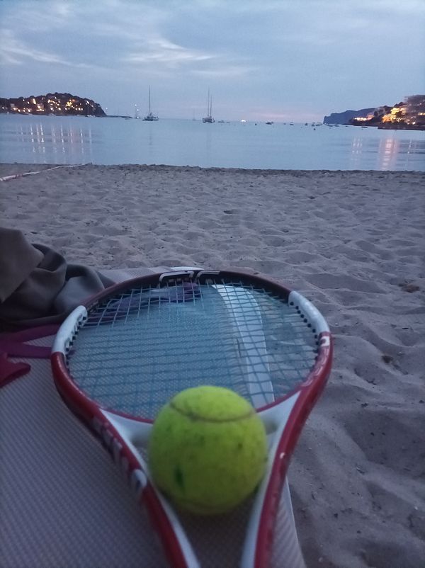 A tennis racket and a ball are positioned on the sandy beach with a serene ocean view at dusk.