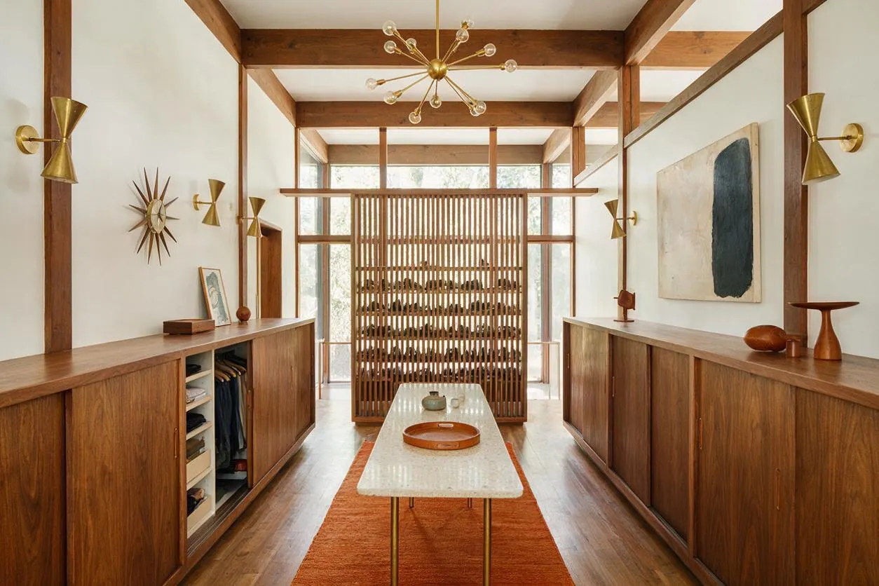 A long, symmetrical closet interior with walnut wood cabinetry on both sides, open shelving and hanging storage, and a central slatted divider at the back. Brass starburst wall sconces and a modern brass chandelier add Mid-Century Modern character, while a pale stone table and rust runner soften the warm wood tones.