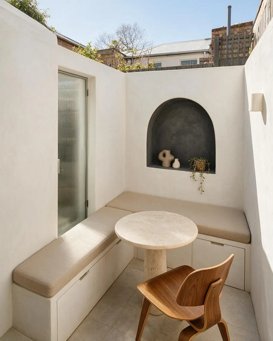 A compact modern backyard seating nook with white stucco walls, a built-in L-shaped bench with beige cushions, and a round stone table. An arched recessed niche with dark plaster displays small ceramics and a trailing plant, while a molded wooden chair and frosted glass door add warmth and texture.