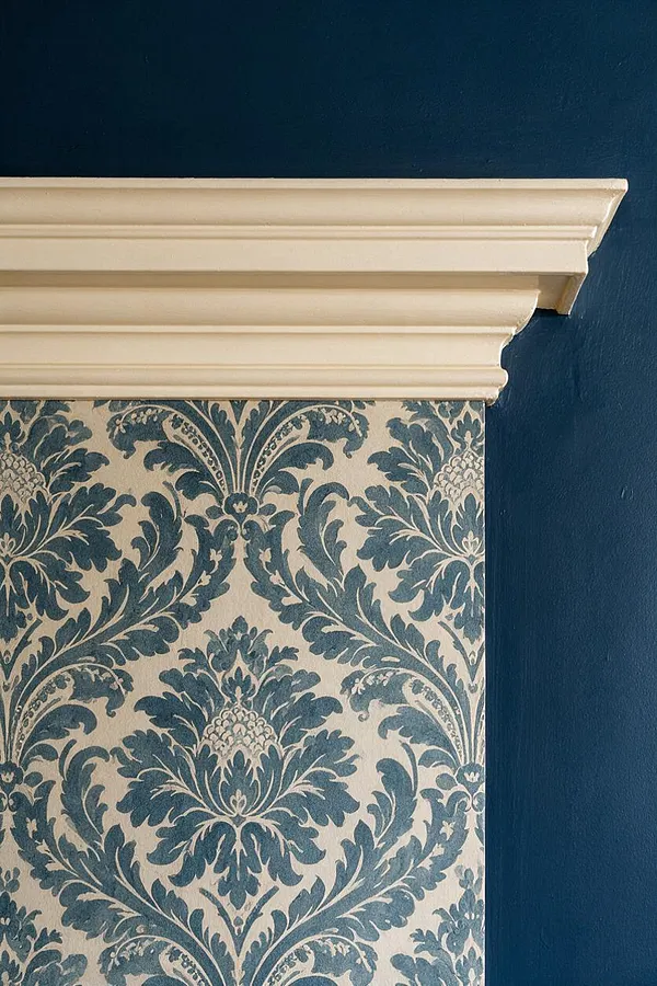 A close-up of a traditional interior wall featuring blue and cream damask wallpaper beneath a cream painted crown molding. The wallpaper has an ornate floral scroll pattern, and the wall above is painted a deep navy blue, creating a rich classic contrast.