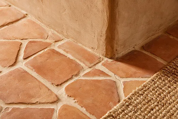 A close-up of a Southwestern interior corner showing irregular terracotta floor tiles with pale grout and natural surface variation. A textured plaster wall meets the warm tiled floor, and a woven natural fiber rug edge appears in the lower right corner.