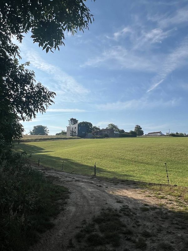A serene landscape in Belgium featuring a grassy field and a few buildings in the distance.