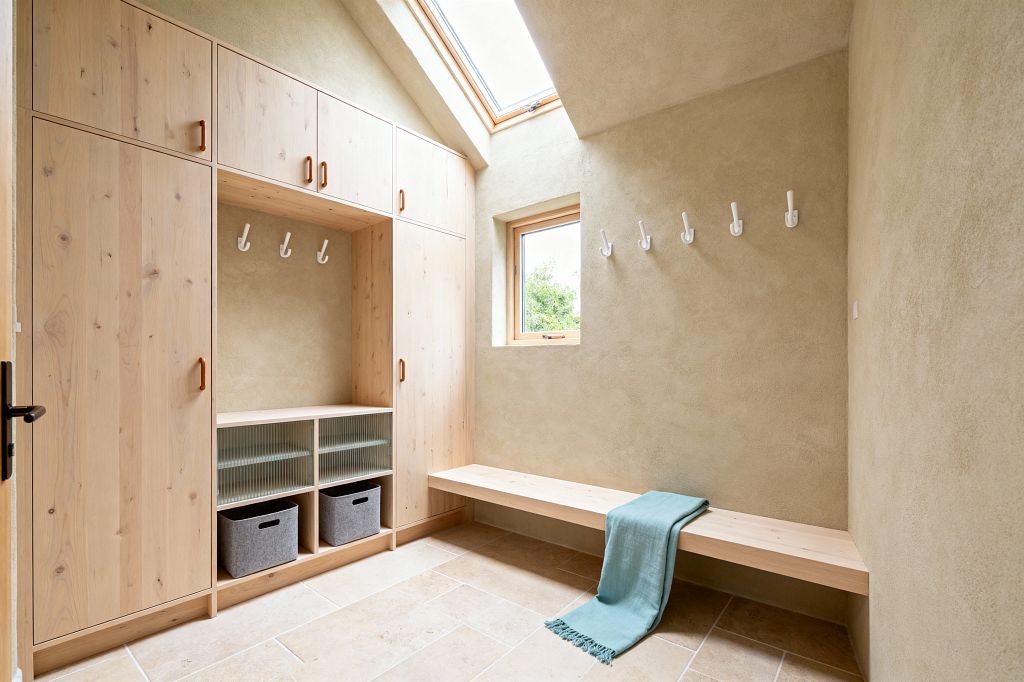 Bright mudroom with built-in pale pine cabinets and a long floating bench. Full-height storage frames an open niche with three white hooks, reeded-glass cubbies, and two gray felt bins. Opposite, a row of white wall hooks sits above the bench. Surfaces are warm beige limewashed plaster and light limestone floor tiles. A small wood-trimmed window and a roof skylight bring in daylight. A muted teal throw drapes over the bench.