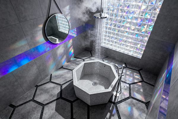 A futuristic gray bathroom shows a ceiling-mounted rain shower pouring into a small octagonal terrazzo soaking tub at the center, with steam rising. The space features hexagonal concrete-look floor tiles divided by black pebble channels, a freestanding stainless steel filler with hand shower, a round strap-hung mirror, and a prismatic glass-block window with an iridescent wall band casting blue and violet reflections.
