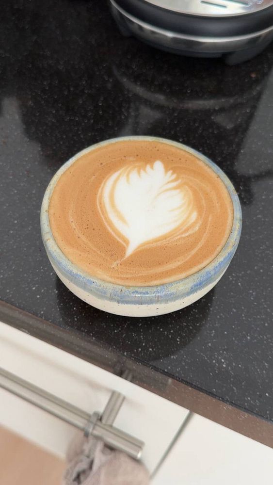 A close-up photo of a cappuccino in a homemade ceramic cup, placed on a white lace surface.