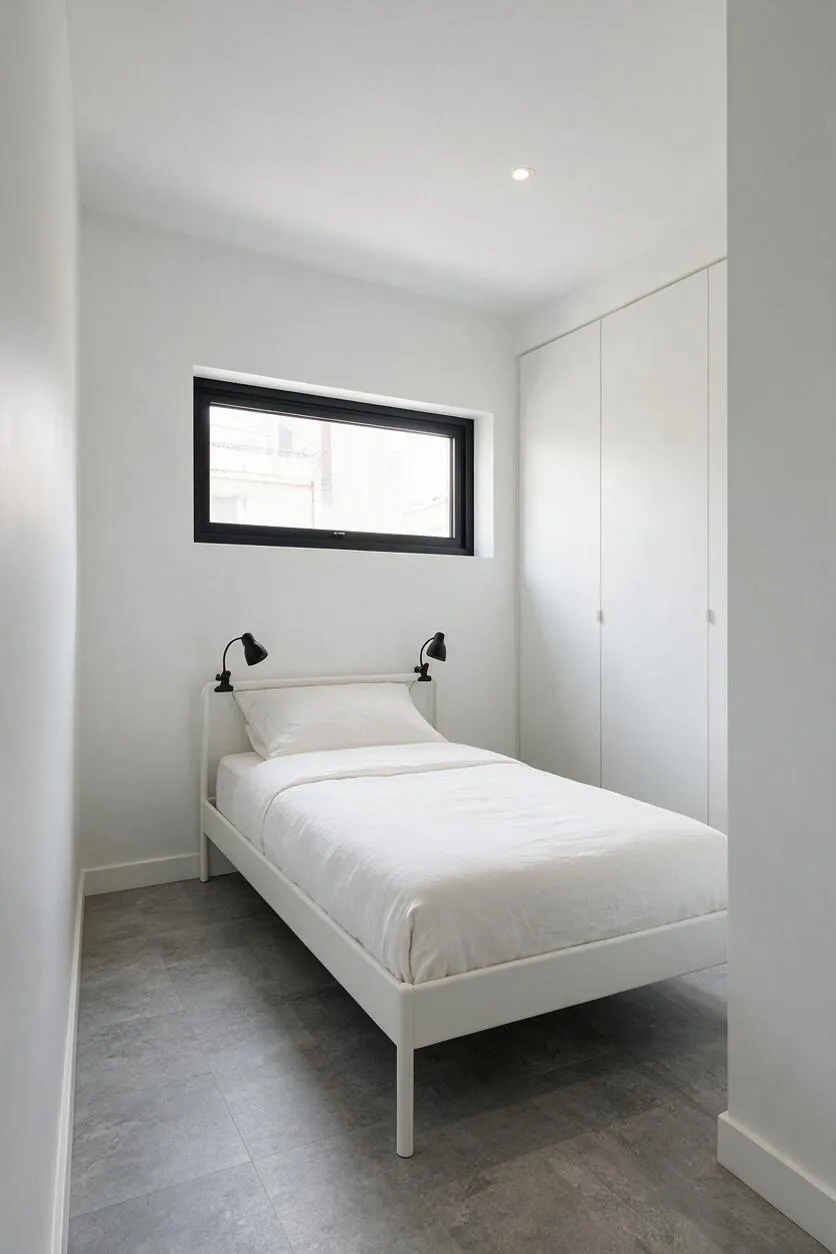 A compact minimalist guest bedroom with a single white metal bed dressed in white bedding, set against white walls. A small horizontal black-framed window sits above the bed, black adjustable reading lamps flank the headboard, and built-in white wardrobes line the right wall. The room has large gray floor tiles and a calm, spare atmosphere.