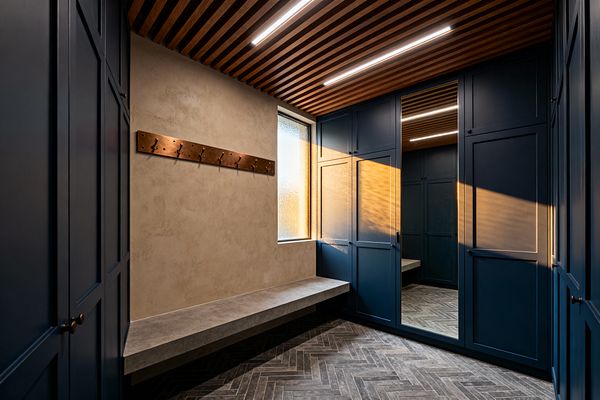 A contemporary mudroom features deep navy built-in cabinets, one with a full-height mirror, framing a corner with a floating gray concrete bench below a frosted window. The room includes a warm wood slat ceiling with linear LED lighting, a brushed copper hook rail on a taupe plaster wall, and charcoal herringbone tile flooring, creating a streamlined, organized entry with rich contrast and softly filtered natural light.