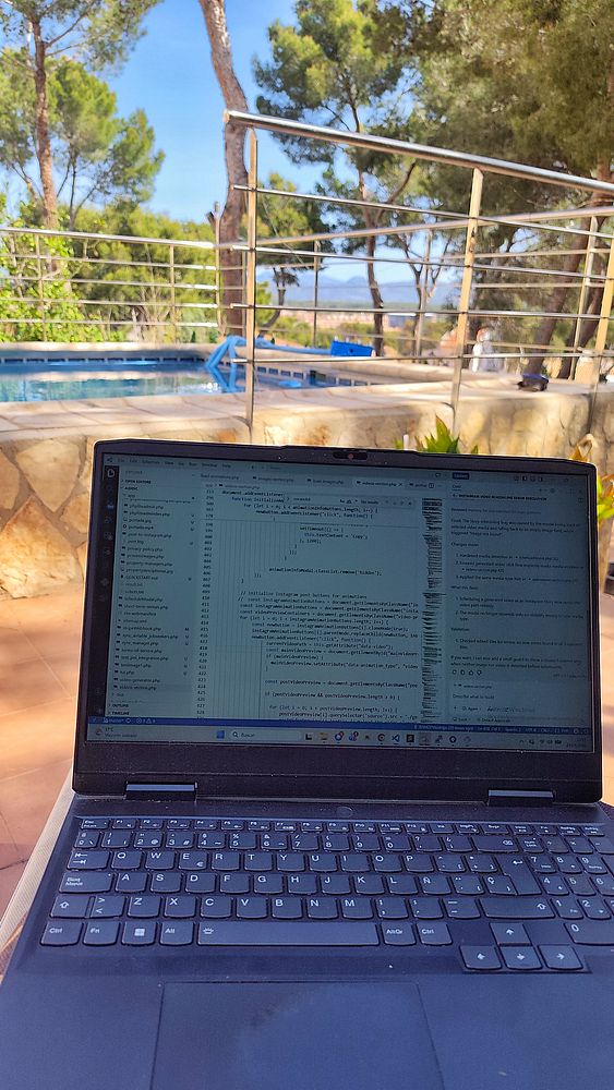 A laptop, smartphone, iced coffee, and eyeglasses are on a wooden table with a pool in the background, indicating a productive work setting by the poolside.