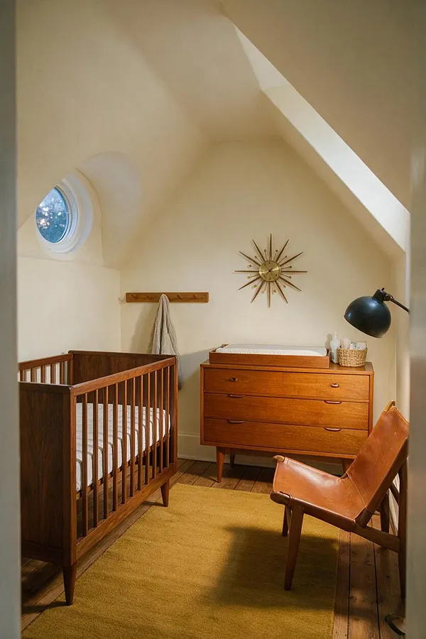 A cozy nursery with a mid-century modern feel features a wooden crib on the left, a teak dresser with a changing tray in the center, and a tan leather chair on the right. Cream walls, a mustard area rug, a sunburst wall clock, a round porthole-style window, and a black task lamp complete the warm, compact room.
