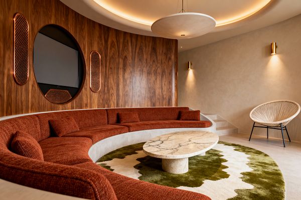 A mid-century modern media room features a curved built-in sofa upholstered in rust fabric around an oval marble coffee table on an olive-and-cream rug. A walnut-paneled wall integrates a circular TV with perforated copper speakers; warm plaster walls, brass sconces, and a saucer-like pendant within a coved ceiling provide soft ambient light, while a natural-cord lounge chair with black legs sits near steps and smooth microcement flooring.