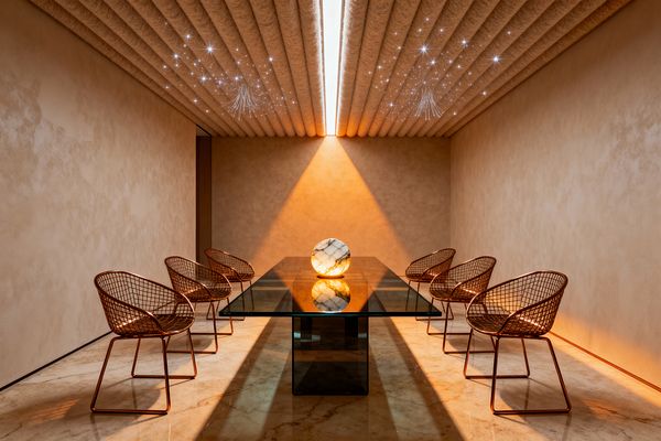 A long, symmetrical dining room features a thick rectangular glass table on a smoked-glass pedestal with six copper-finish wire chairs, three on each side. Warm beige plaster walls and polished cream marble flooring glow under a central linear ceiling light and a star-like array of fiber-optic points in ribbed felt panels; a luminous marble sphere sits centered on the table, enhancing the minimalist, futuristic atmosphere.