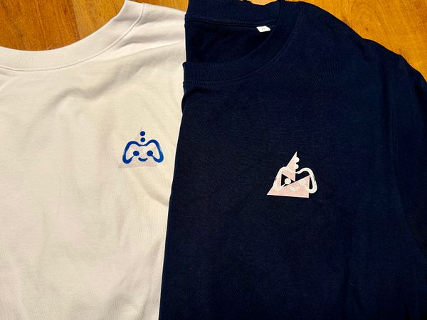 Three white T-shirts with colorful illustrated cards pinned to each are displayed on a beige surface.