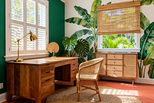 A bright tropical home office with a solid wood desk, brass task lamp, and a small woven fan sculpture sits beside white plantation shutters. A rattan chair rests on a round jute rug. The adjacent wall displays a large banana-leaf and monstera mural next to a window with a bamboo Roman shade, above a cane-front sideboard. Sunlight and lush greenery create an airy, natural look.