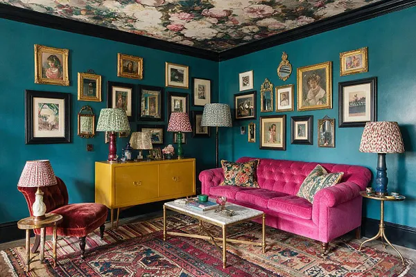 A vivid studio apartment living area with teal walls, a floral-painted ceiling, and a gallery of framed artwork in mixed black and gold frames. A hot pink tufted sofa, burgundy velvet chair, mustard cabinet, patterned lamps, layered rugs, and brass tables create a richly textured maximalist look.