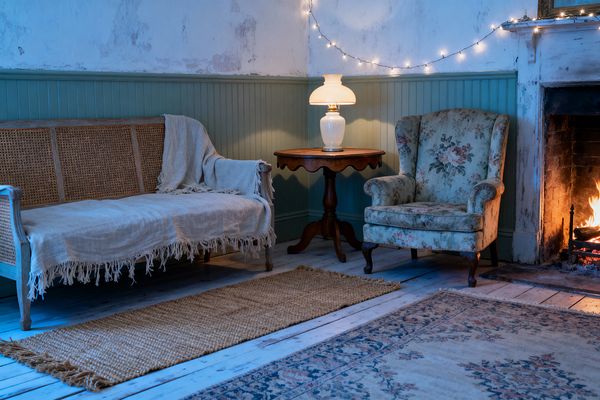 A shabby chic living room with weathered plaster walls above sage-green beadboard features a cane-back wooden settee draped in a fringed linen throw, a round dark wood pedestal table with a white milk-glass lamp, and a floral upholstered wingback chair beside a lit fireplace. Distressed white floorboards hold a jute runner and a faded patterned rug, while string fairy lights add a soft, warm glow across the wall.