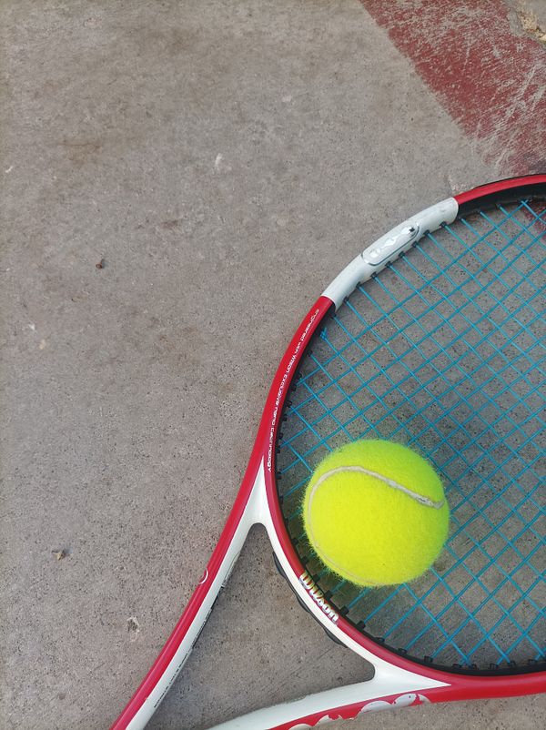 A tennis racket and a bright yellow tennis ball are positioned on a concrete surface.