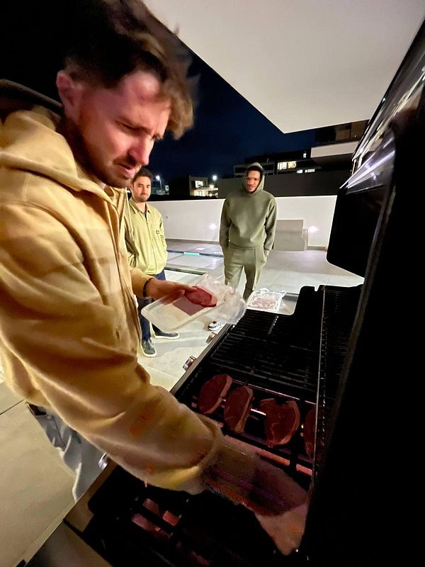 A group of three men is gathered around a grill at night, with one man preparing to cook steaks.