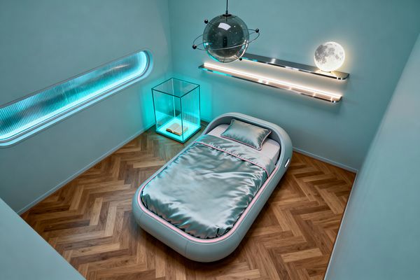 A compact kids' bedroom with soft aqua walls features a rounded pod-style single bed dressed in metallic aqua satin with pink piping on warm herringbone wood floors. Overhead hangs a smoked-glass planet pendant; a floating chrome shelf with LED strips and a glowing moon lamp lines one wall, while a backlit ribbed window and a cyan-lit glass cube side table add futuristic highlights in a tidy, space-themed setting.
