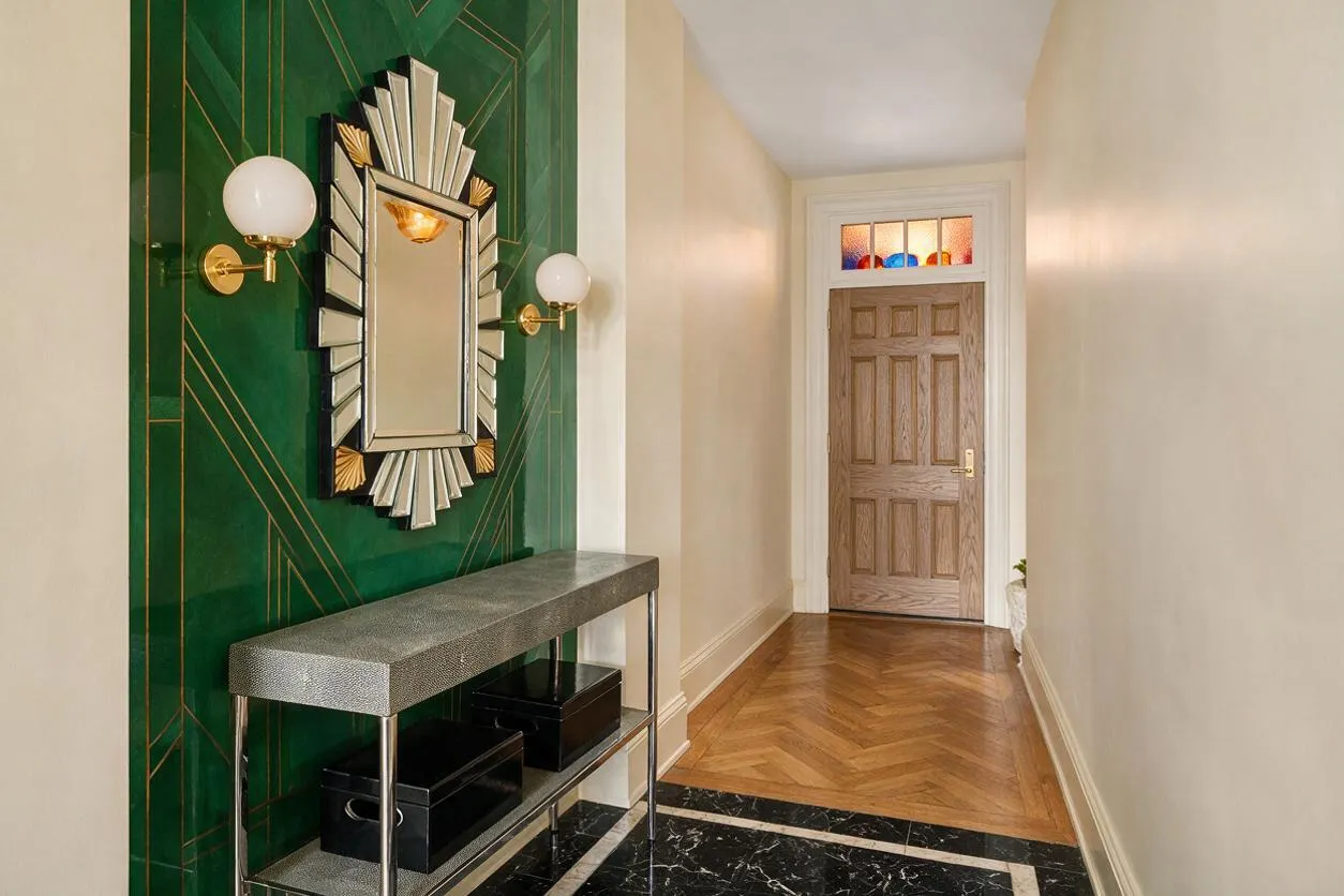 A narrow entryway with an emerald green Art Deco accent wall on the left, featuring gold geometric linework, a sunburst-style mirror, and brass globe sconces. A slim console table with a textured gray top and black storage boxes sits below, while the hallway leads to a wood front door with stained glass transom and warm herringbone flooring.