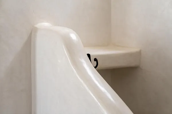 A close-up of a Moroccan-style staircase finished in smooth glossy white plaster, with a curved built-in handrail or stair edge and a soft beige plaster wall and landing behind it. A small black wrought-iron detail peeks from the side, adding contrast to the minimalist organic form.