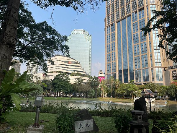 A serene urban park scene featuring a pond surrounded by modern buildings and lush greenery.