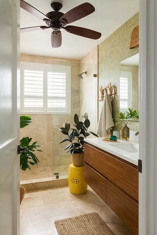 A tropical-style master bathroom with a dark wood ceiling fan, beige stone-tile walls, and a glass walk-in shower with white plantation shutters above it. A floating wood vanity with a white countertop runs along the right side, paired with a large mirror, woven wall decor, and a taupe towel. Potted tropical plants and woven accessories add a relaxed, resort-like feel.
