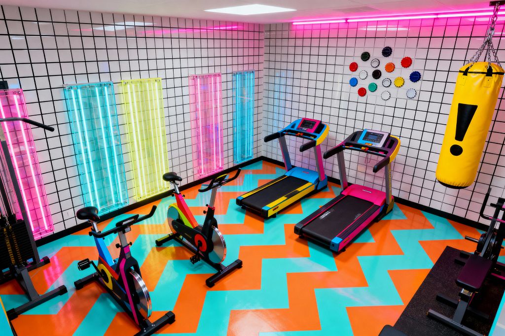 A vividly colored home gym with white square tile walls grouted in black, vertical neon panels in pink, cyan, and yellow, and a pink LED strip along the ceiling. A glossy teal and orange zigzag epoxy floor anchors two color‑block spin bikes, two treadmills with cyan, magenta, and yellow accents, and a yellow hanging punching bag with a black exclamation mark. A cable machine and a weight bench sit at the sides on rubber mats.