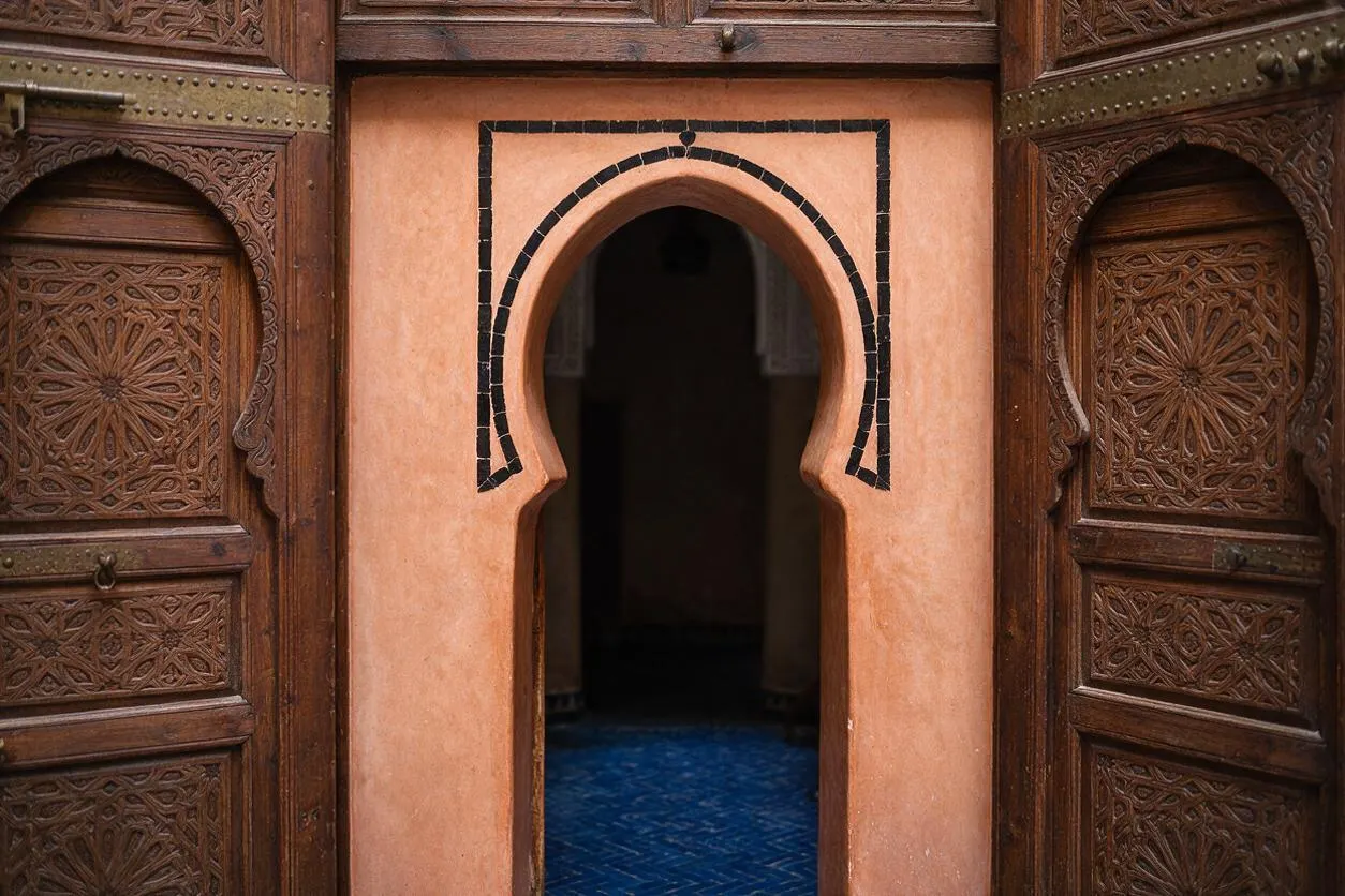 A Moroccan-style exterior entrance features richly carved wooden doors on both sides of a peach-toned stucco wall. A central horseshoe arch outlined in dark tile opens into a dim interior with a blue patterned floor.