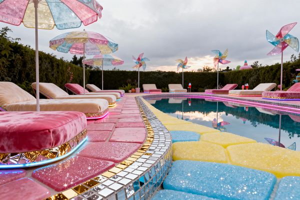 A curved swimming pool is bordered by candy-colored pavers that sparkle in pink, yellow, and sky blue, edged with mirrored mosaic and gold tiles. Along the deck, plush cream and pink lounge chairs rest on glitter-trim bases with LED underlighting and are shaded by pastel patchwork umbrellas. Oversize translucent pinwheels rise behind the seating, with tall hedges enclosing the patio at dusk. The water reflects the lights and colors, emphasizing the playful, candy-inspired design.