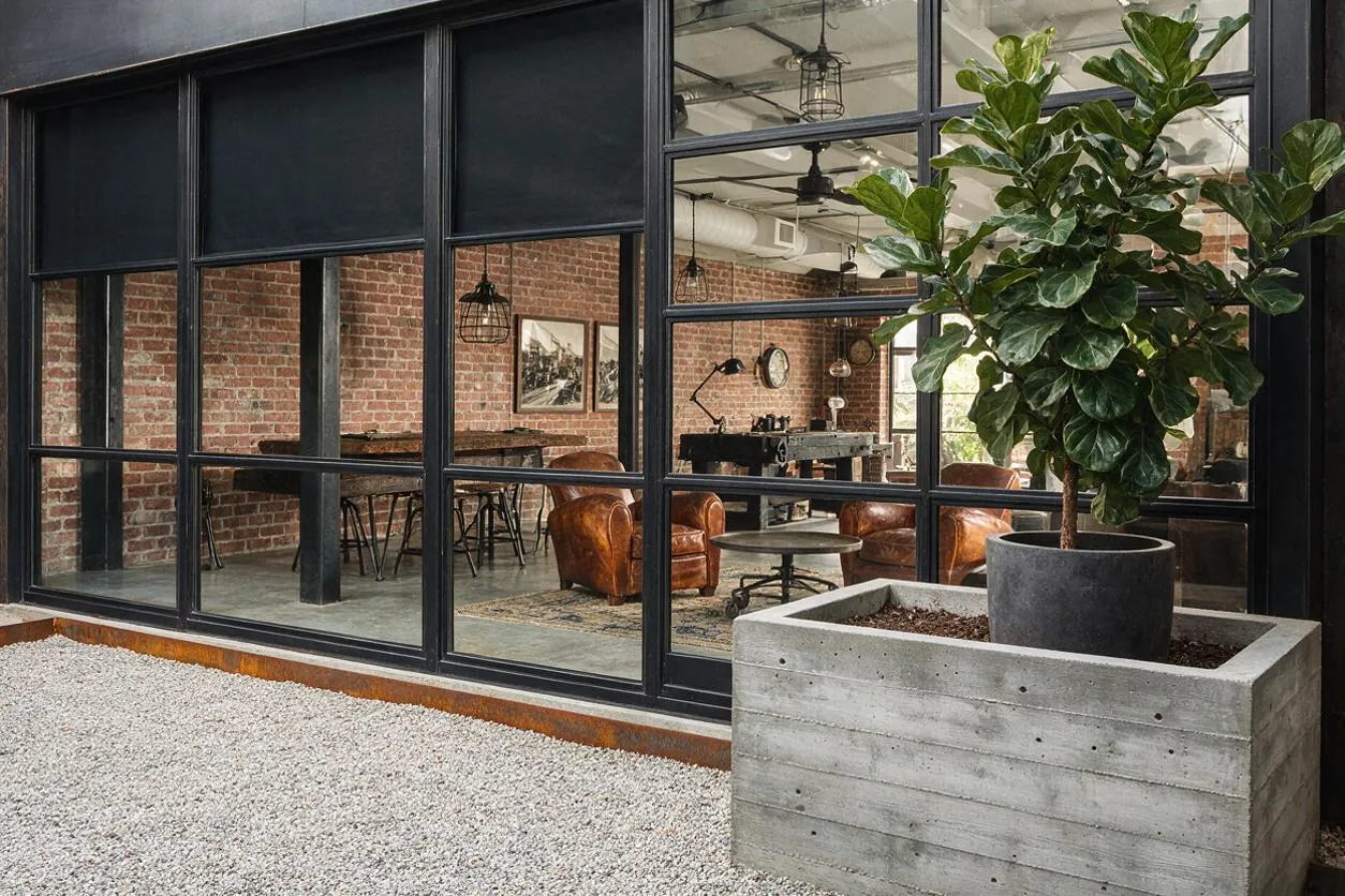 An industrial-style exterior with large black steel-framed glass windows and doors revealing a brick-walled interior. A concrete planter with a leafy tree sits on a gravel forecourt, reinforcing the raw, modern material palette.