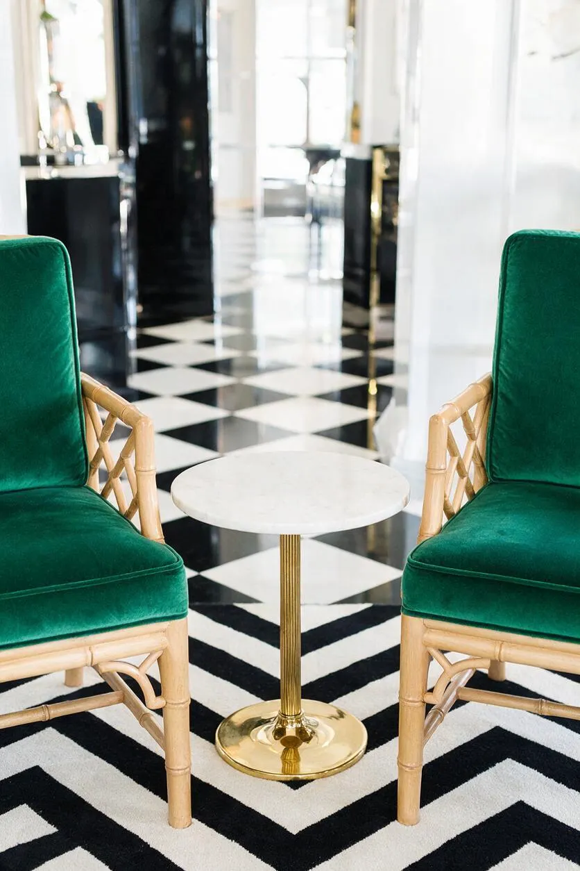 A glamorous seating vignette with two emerald green velvet chairs facing a small round white tabletop on a polished brass pedestal. The room features a bold black-and-white geometric floor and bright white walls, creating a refined Hollywood Regency look.