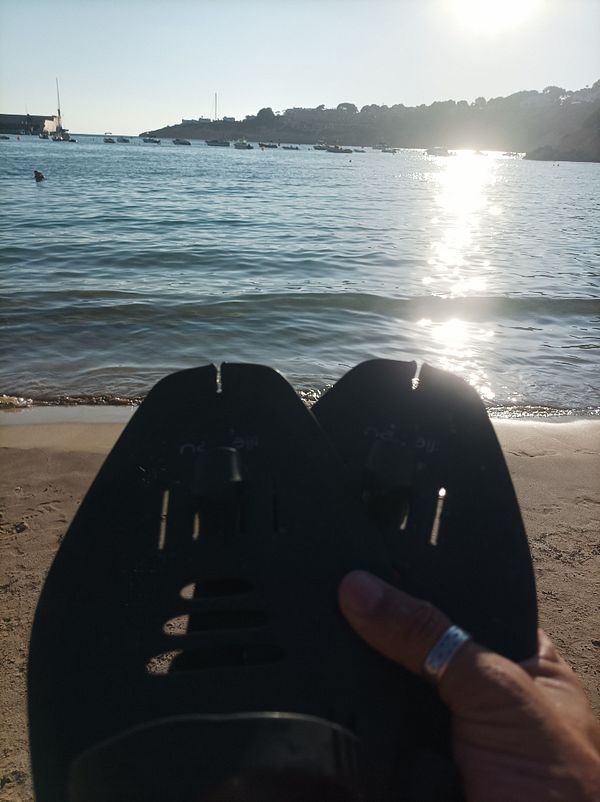 A person holds swim fins while sitting on the beach, with a scenic view of the water and boats in the background.