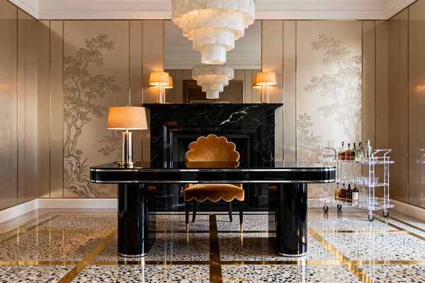 A luxurious home office centers on a glossy black lacquer desk facing a black marble fireplace, flanked by warm brass lamps and an alabaster tiered chandelier above. Champagne metallic wall panels with chinoiserie trees, a caramel velvet scalloped chair, a clear lucite bar cart, and terrazzo flooring with brass inlay create a vintage glam palette, softly lit for an intimate, polished atmosphere.