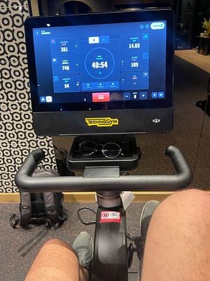 A person is exercising on a Technogym stationary bike, with a digital display showing workout metrics.