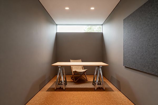A compact minimalist home office shows a light wood trestle desk centered on a cork floor with a beige canvas sling chair behind it. Warm gray walls and a soft white ceiling enclose the narrow room, illuminated by a slim horizontal clerestory window and two recessed ceiling lights. A large gray felt panel hangs on the right wall, adding acoustic control and texture while the uncluttered surfaces and muted palette create a calm, focused work setting.