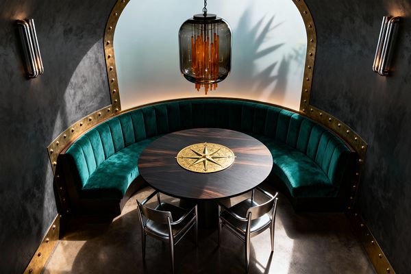 A steampunk-inspired dining nook features a curved emerald velvet banquette set within a riveted brass arch against charcoal plaster walls, surrounding a round dark walnut table inlaid with a large brass compass rose and paired with two brushed metal chairs. Overhead, a smoked-glass pendant with cascading amber tubes glows before a backlit frosted window, with streamlined metal sconces and a polished concrete floor completing the scene.