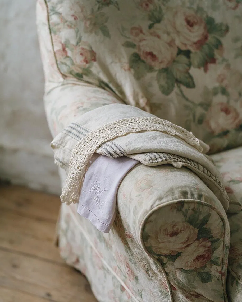 A close-up of a cushioned armchair upholstered in faded floral fabric with cream, pink, and green tones. A folded linen cloth with striped trim and a crocheted lace edge is draped over the rounded arm, creating a soft cottagecore look.