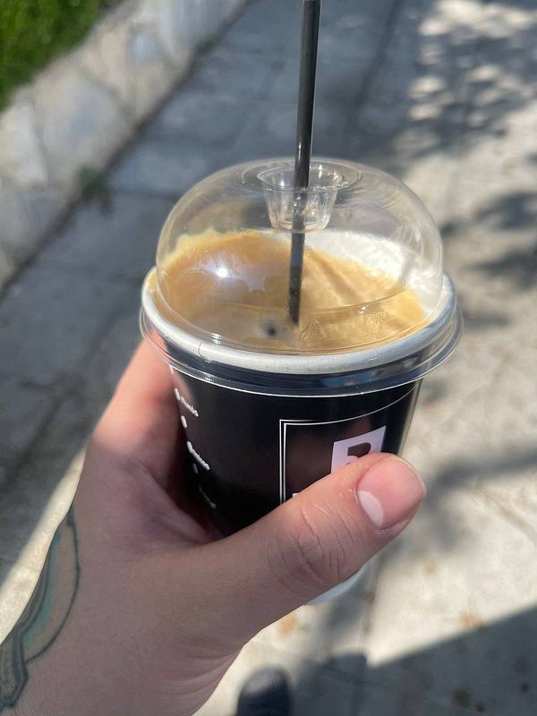 A hand holds a coffee cup while walking on a sunny sidewalk.