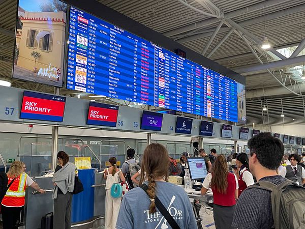 A busy airport check-in area with a large flight information display and travelers waiting in line.