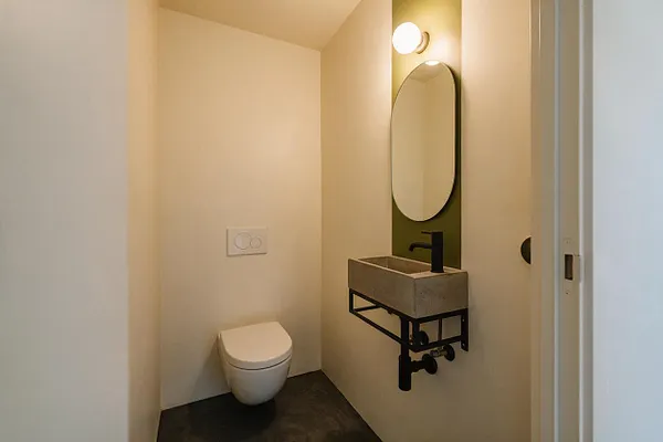 A small modern powder room with a wall-mounted white toilet on the left, a rectangular concrete sink on a black metal frame, and an oval mirror above it. Warm beige walls, a dark floor, and a single globe light create a minimalist, softly lit space.